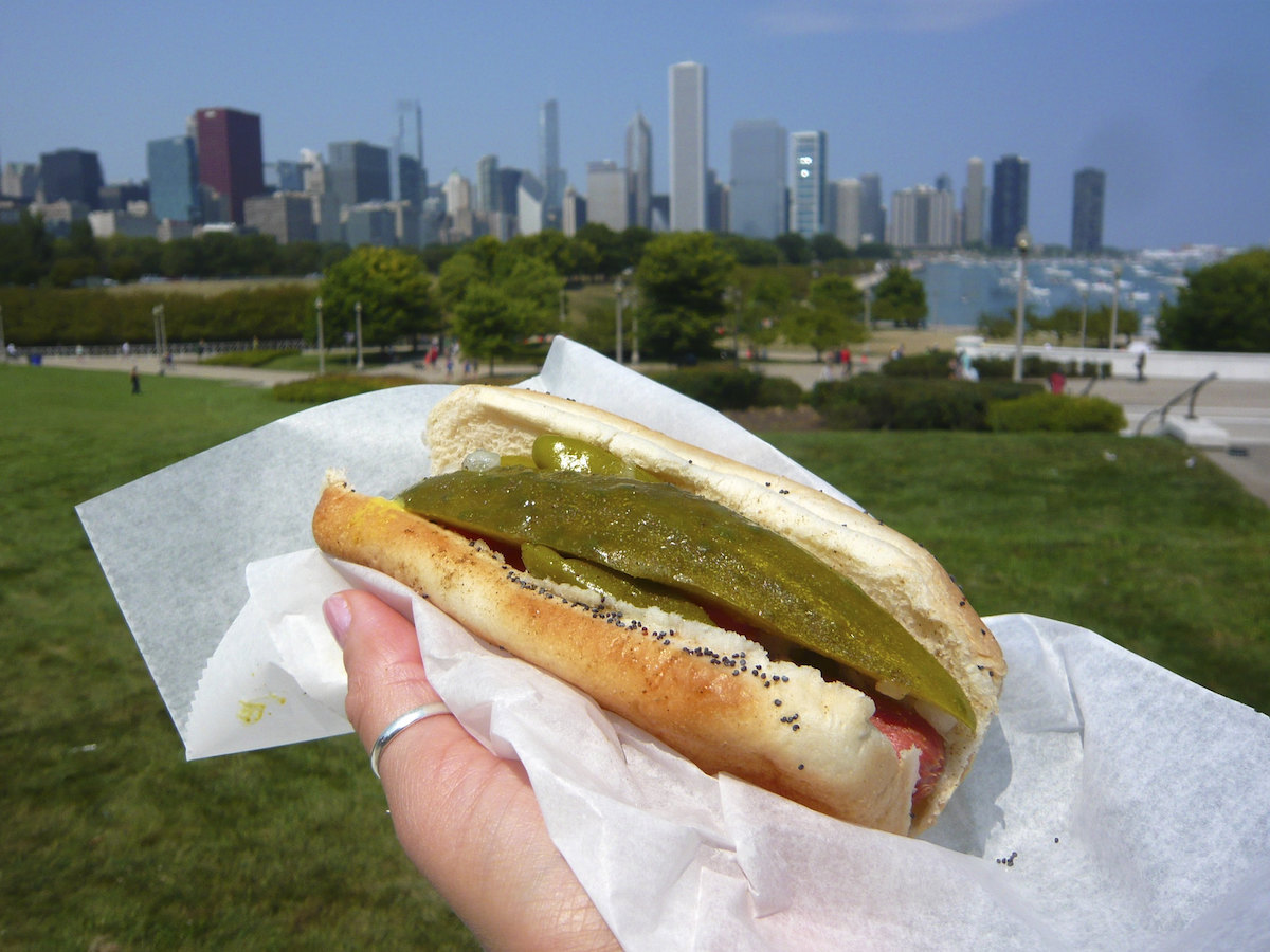 a person holds a Chicago-style hotdog with a polish sausage and pickle spears in a bun with the Chicago skyline in the background