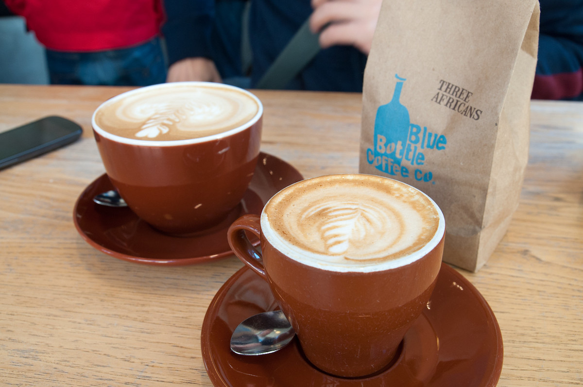Close up of two cappuccinos in brown cups with a paper bag with blue writing that reads "Blue Bottle Coffee"