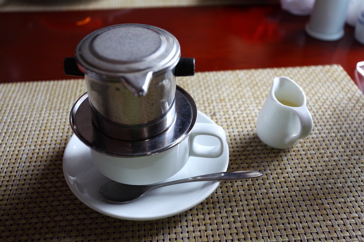 close up of a white coffee cup and saucer with a metal drip filter device over the cup, making Vietnamese style coffee