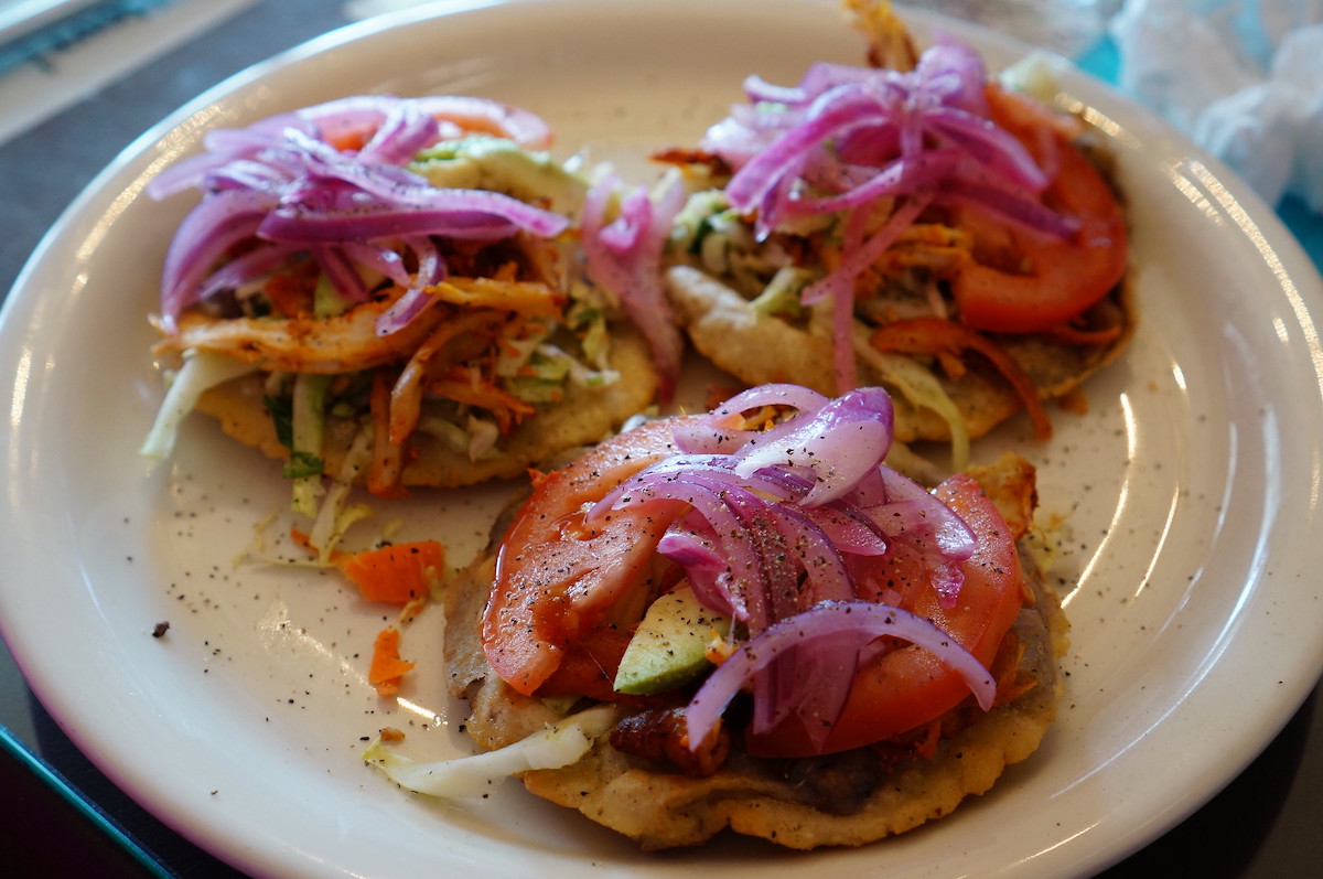 close up of a plate with three panuchos, flat fried tortillas with toppics of tomato, cabbage, avocado, and red onion