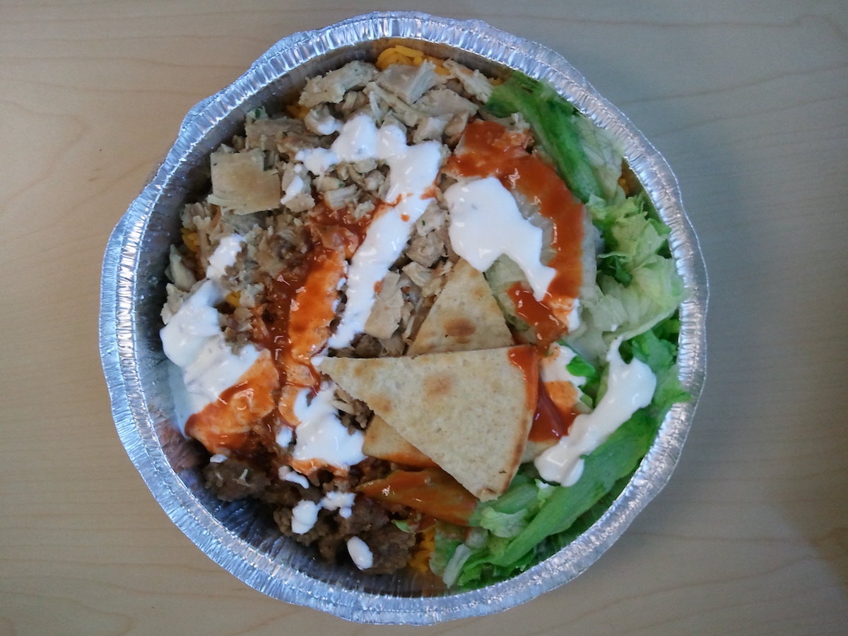 A round aluminum container filled with cooked chicken, rice, lettuce, and pita all topped with drizzles of red and white sauce made by Chicken & Rice Guys, a street food vendor in Boston