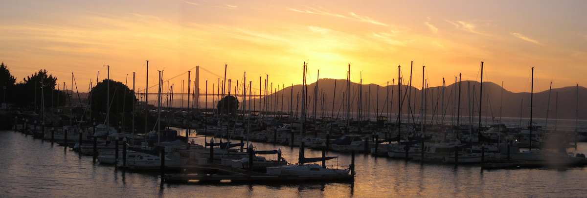 Docked sailboats lined up on the water of the San Francisco Bay with the sunsetting behind the Golden Gate Bridge in the background