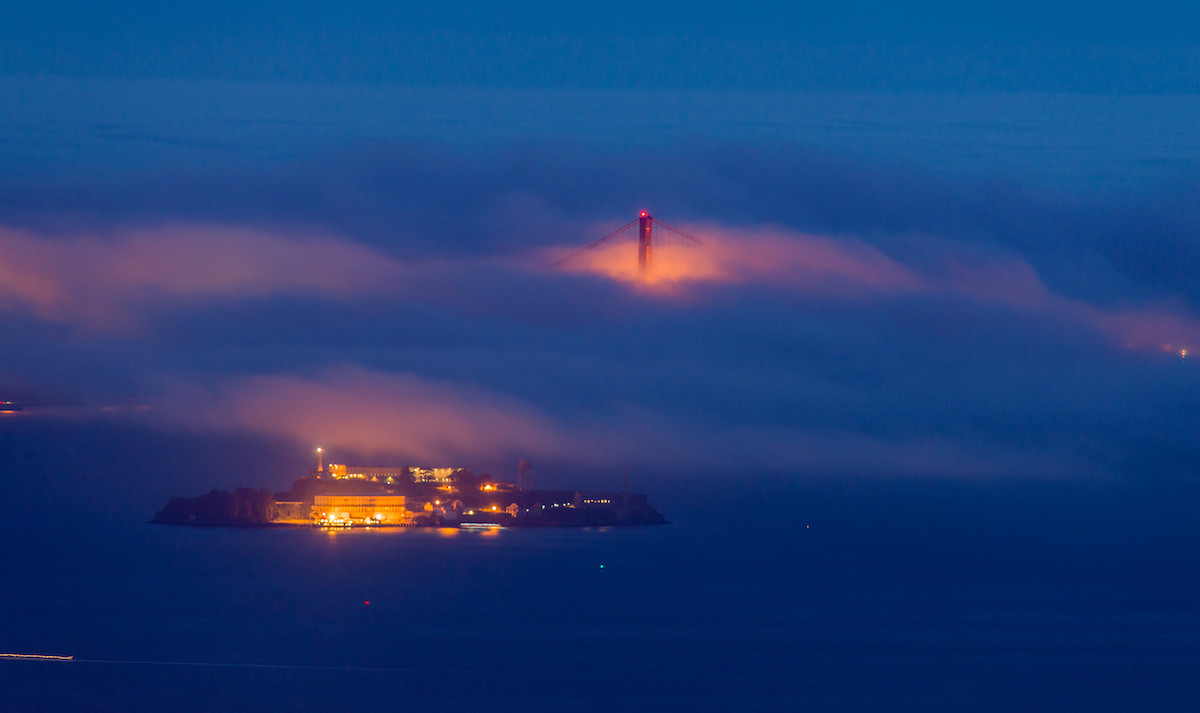 A photo of Alcatraz Island in San Francisco at night, with the top of the Golden Gate Bridge peeking out from the fog