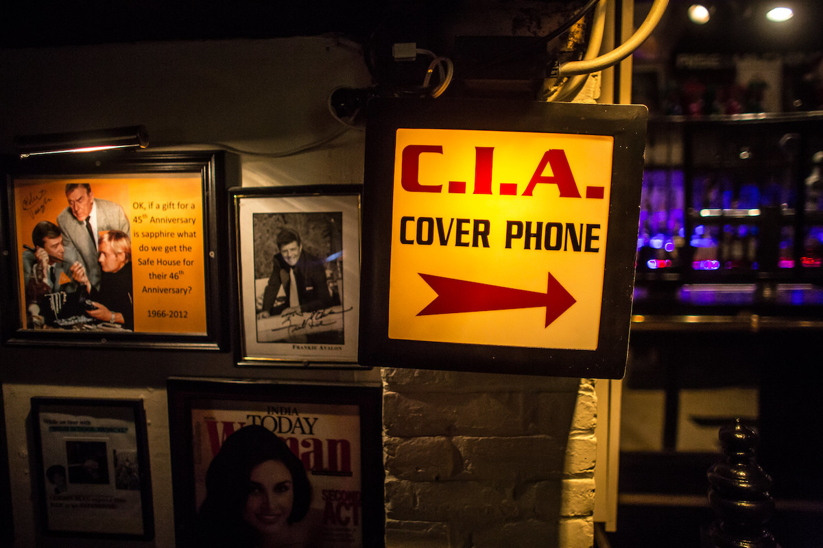 A wall in a spy-themed restaurant covered in pictures and notes, with a big illuminated sign that reads "C.I.A. Cover Phone" 