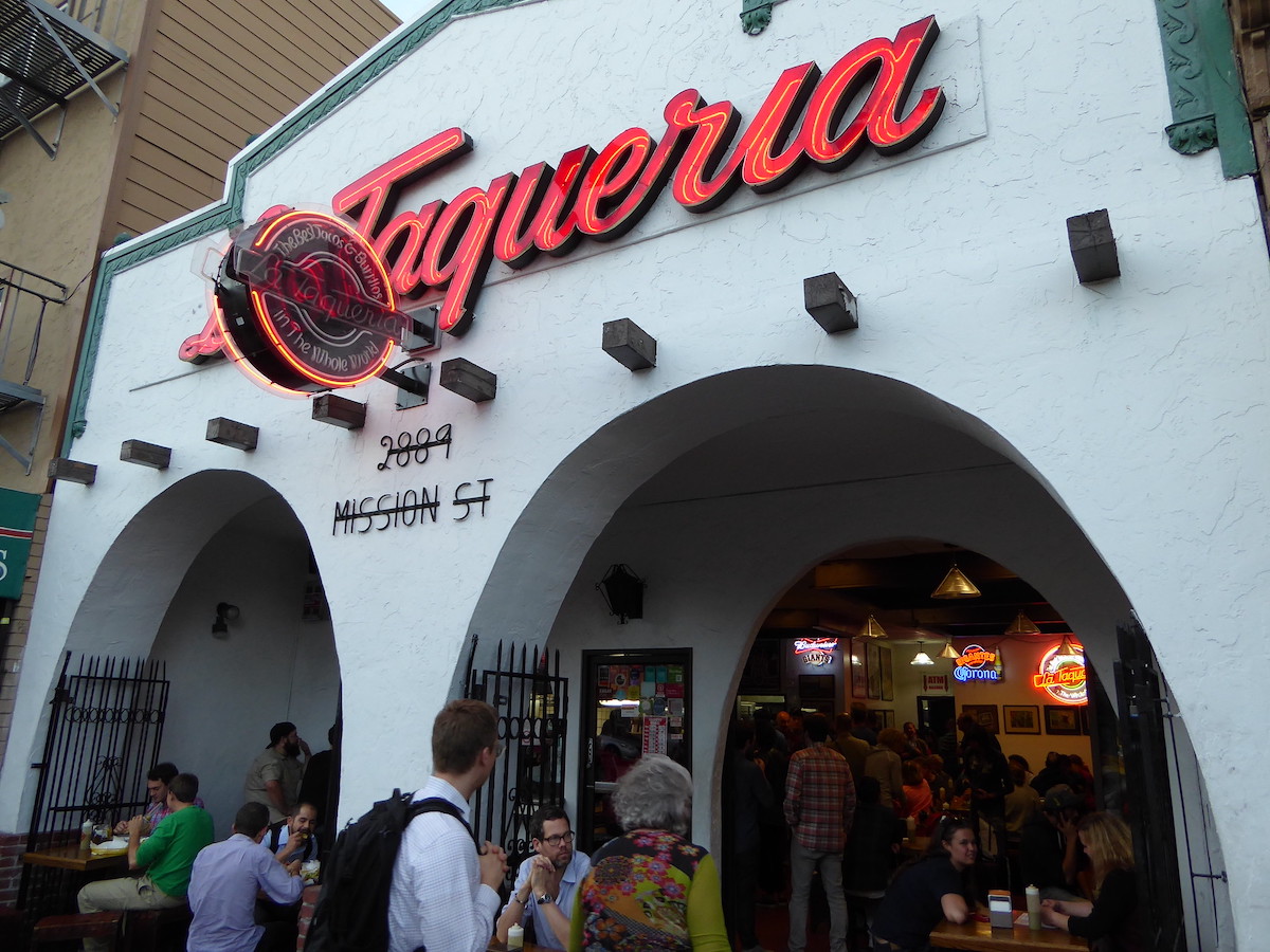 Mexican restaurant with white-painted facade, two arches, and a big red neon sign that reads "La Taqueria"