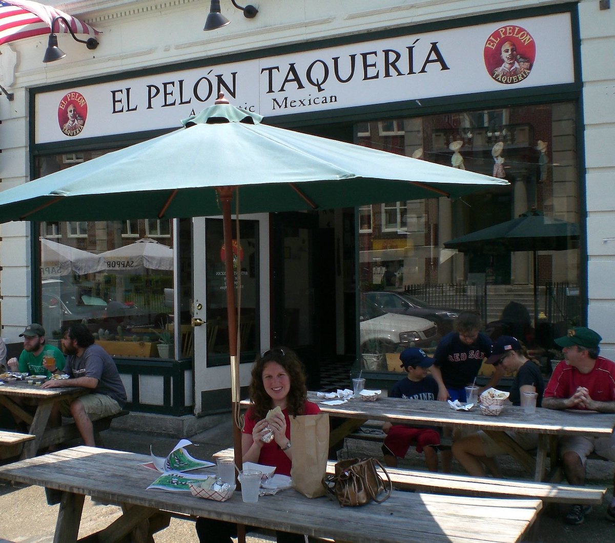 Picture of the exterior of a restaurant in Boston with picnic tables and green umbrellas. Over the door, a white sign with black letters reads "EL PELON TAQUERIA"