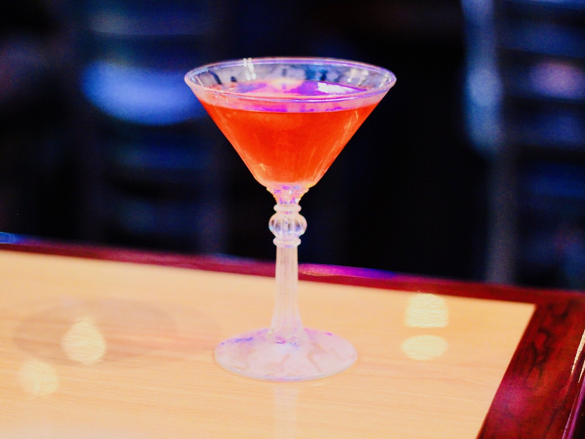 Close up of a bright red cocktail in a martini glass sitting atop a wooden bar