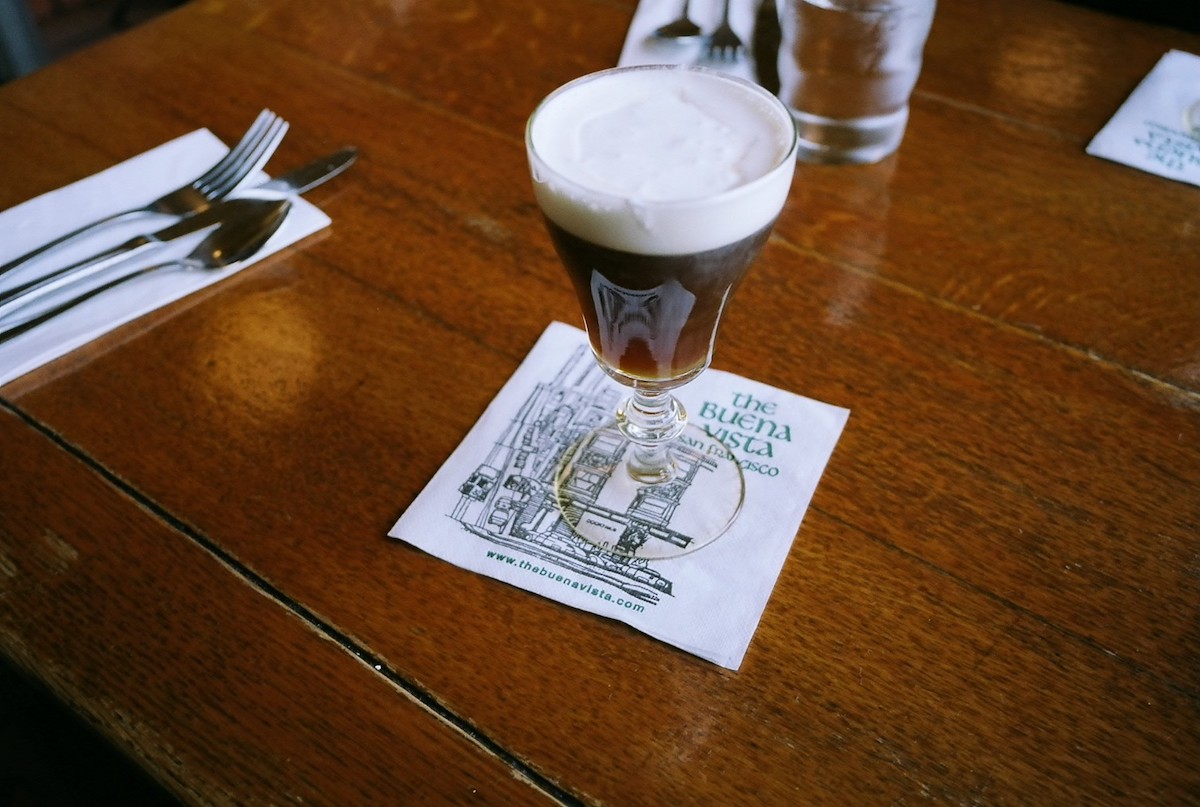 A polished wooden table with a clear goblet of foamy Irish coffee, atop a white napkin that reads "The Buena Vista"