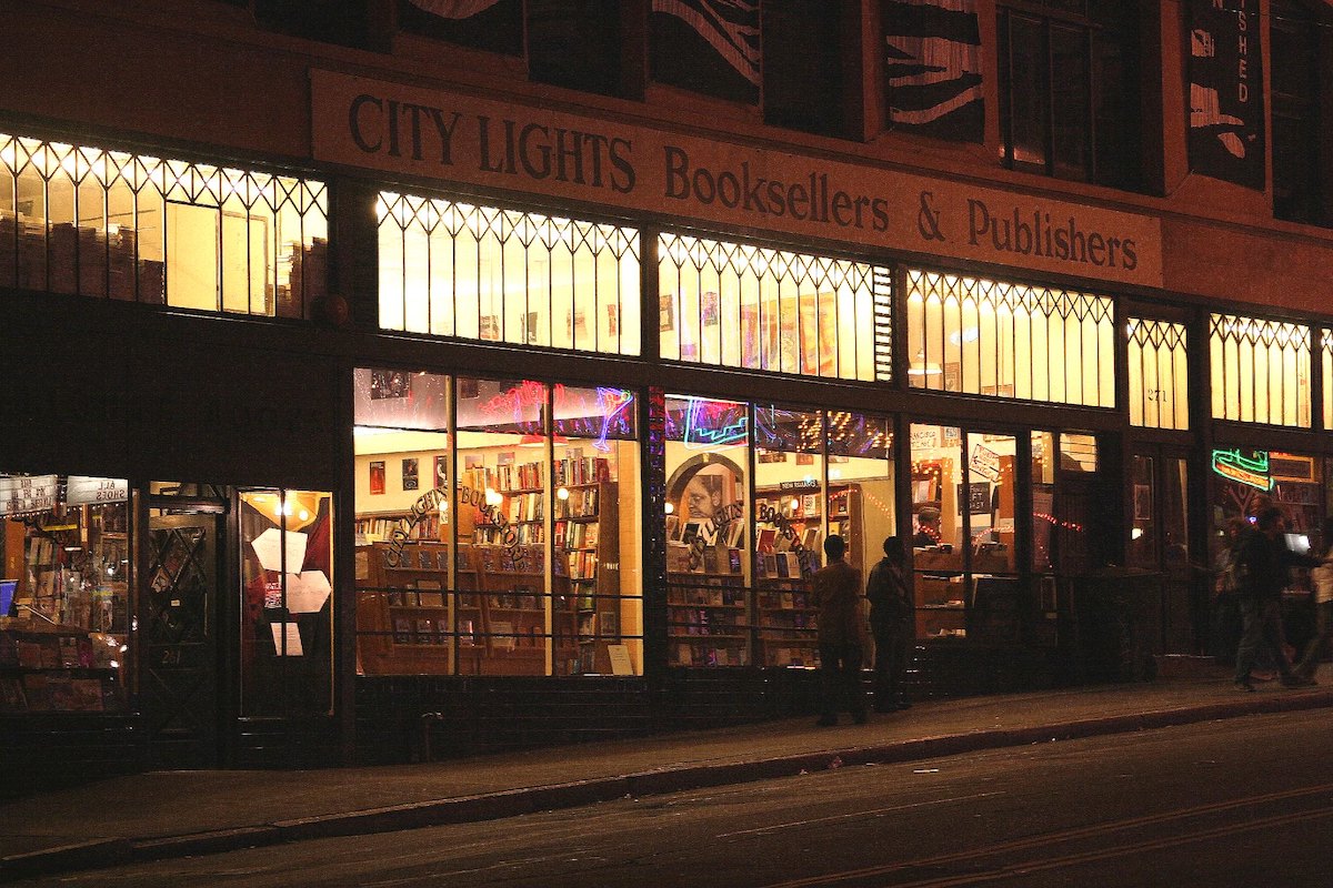 Exterior shot of the City Lights Booksellers & Publishers in San Francisco. It's nighttime and the interior is all lit up, so we can see the bookselves through the windows