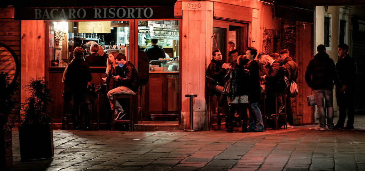A shot of a corner tavern at night in Venice, lit up by red lights. People sit and stand at tables, enjoying wine and snacks.