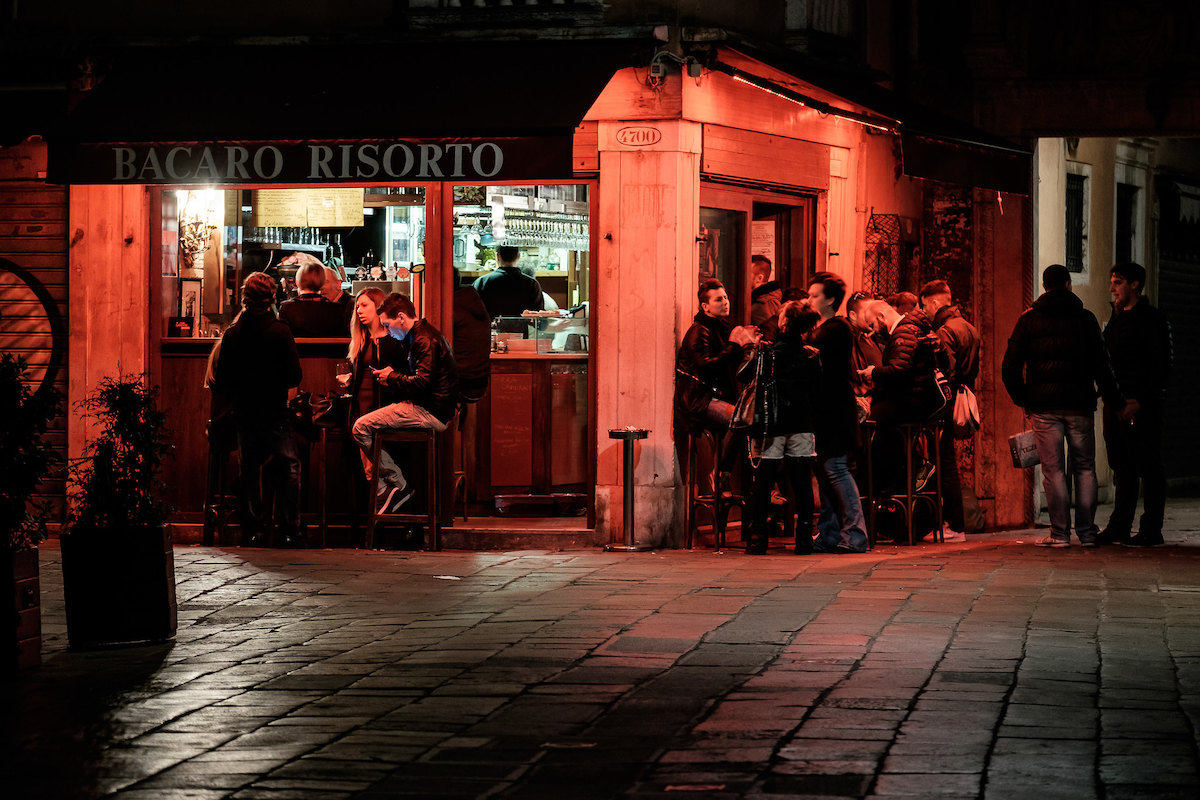 A shot of a corner tavern at night in Venice, lit up by red lights. People sit and stand at tables, enjoying wine and snacks.