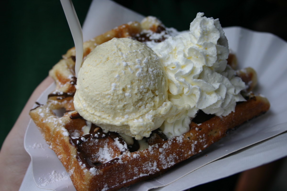 close up of a paper tray with a small Belgian waffle topped with chocolate syrup, a scoop of gelato, and whipped cream