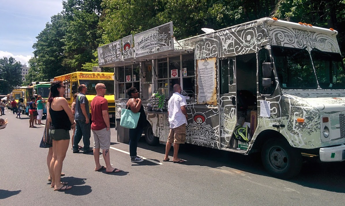 People line up to order at a food truck at the Boston food truck festival. The truck is parked on the street with dozens of other food trucks parked in a line fading into the background.
