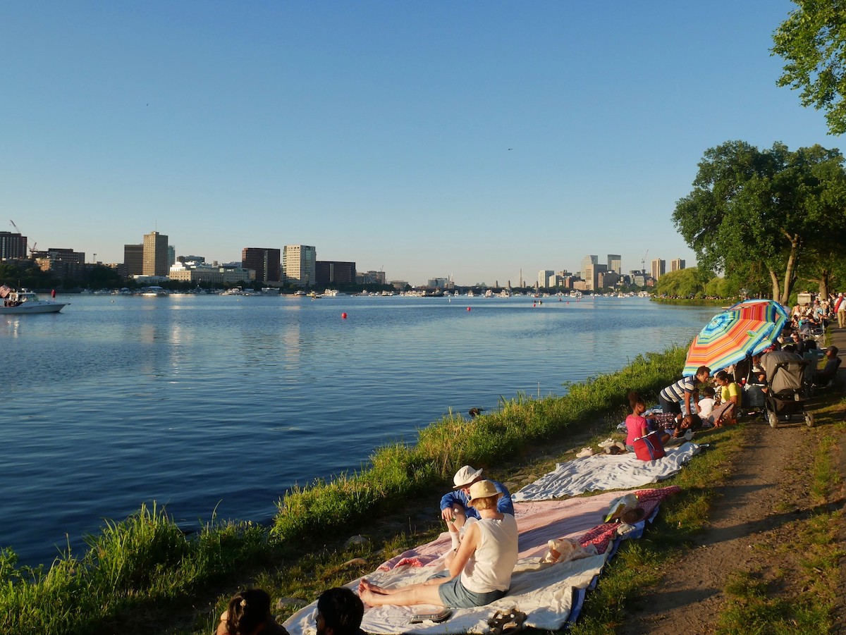 Lots of people line the bank of the Charles River sitting on blankets and enjoying a great July 4th picnic spot in Boston