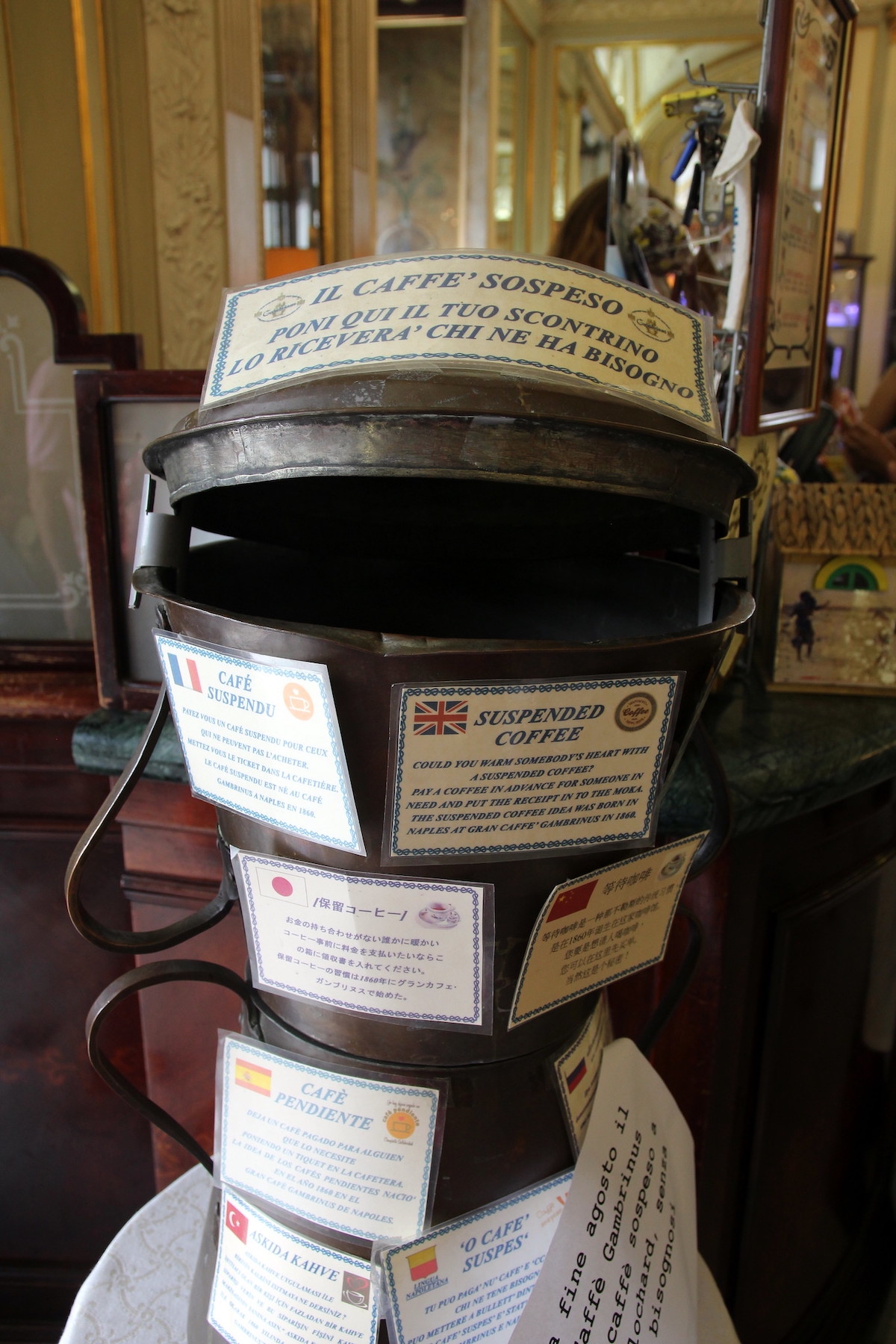 A old coffee pot is covered with cards for "caffe sospeso", a tradition where a patrom of a coffee shop can purchase an extra coffee and leave behind a receipt so that someone down on their luck can come and enjoy a coffee for free