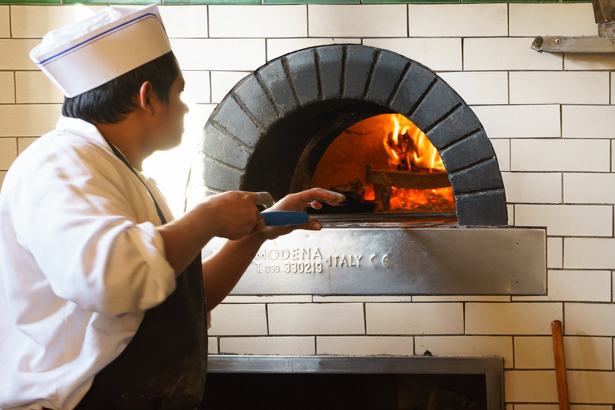 A chef whereing a white jacket and apron uses a pizza peel to bake a pizza in an authentic wood fire pizza oven from Italy