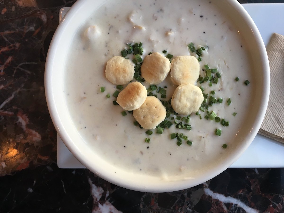 Flat lay shot of a bowl of New England clam chowder topped with chives and oyster crackers on a black marble table