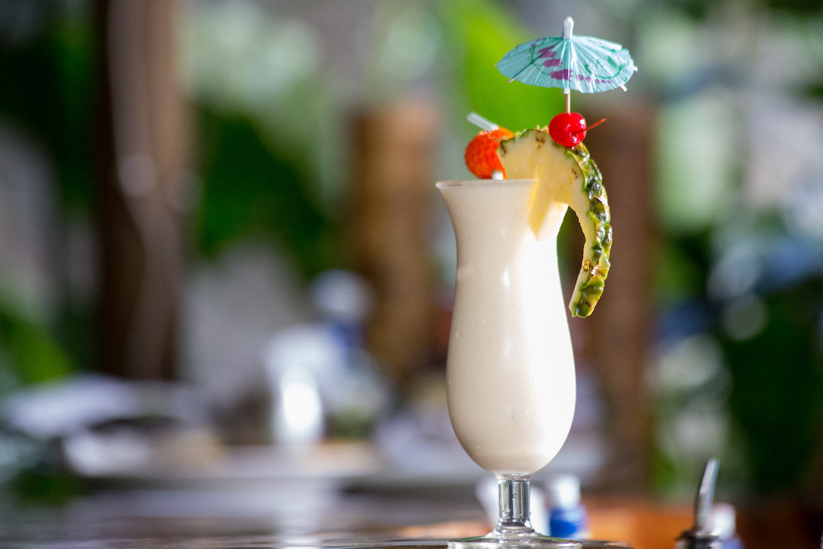 A piña colada cocktail in a hurricane glass witha pineapple slice and cherry garnish sits on a bar with tiki themed decorations in the background