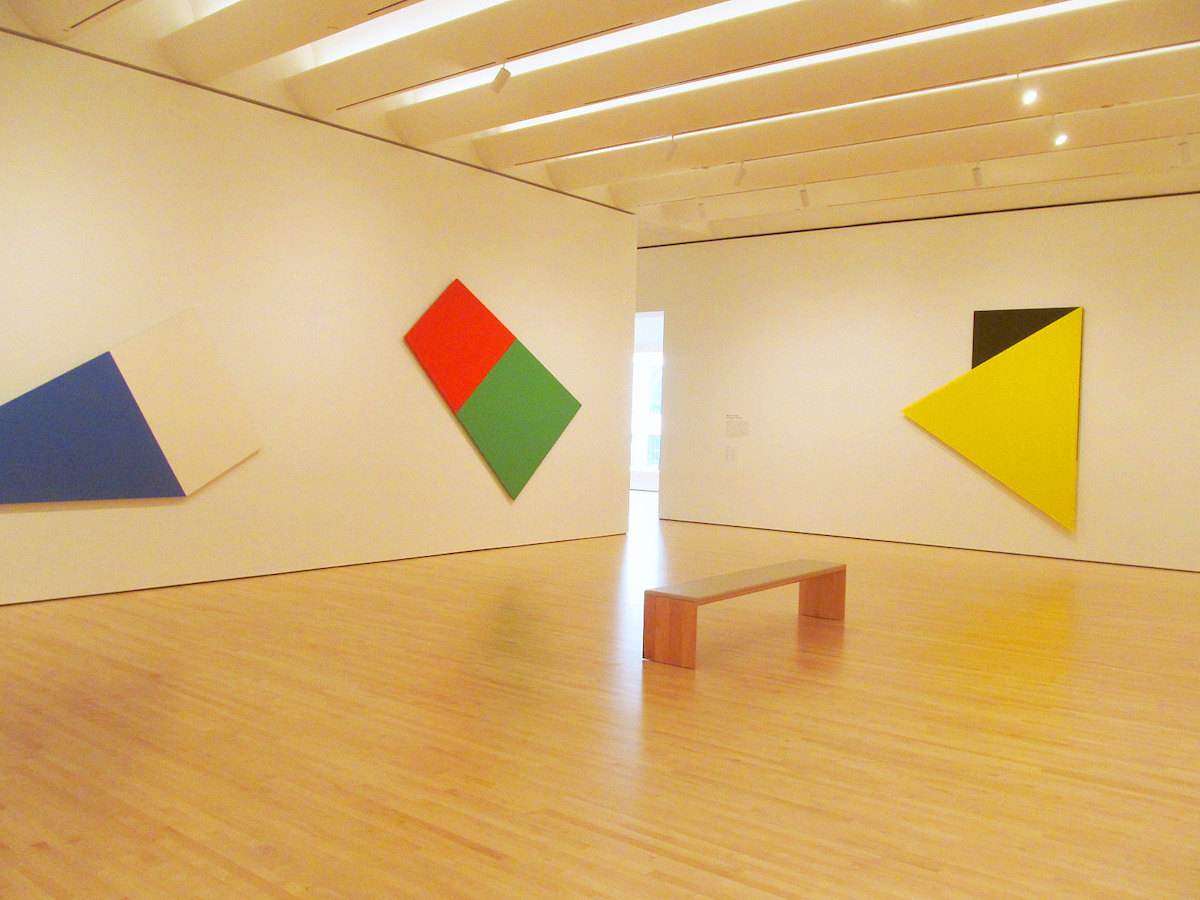 Interior shot of a gallery in SFMOMA. The room has white walls and wooden floors, with a wooden bench in the middle of the room and abstract art on the walls made up of brightly colored shapes.