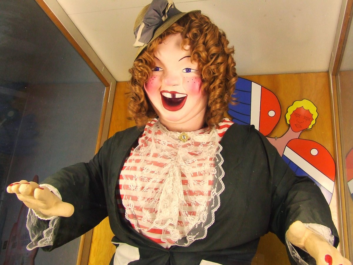 Close up of an animatronic character named Laffing Sal. She is dressed in early 1900s clothes with a hat perched on the side of her head. She has red curly hair, rosy cheeks, and a big open-mouth smile