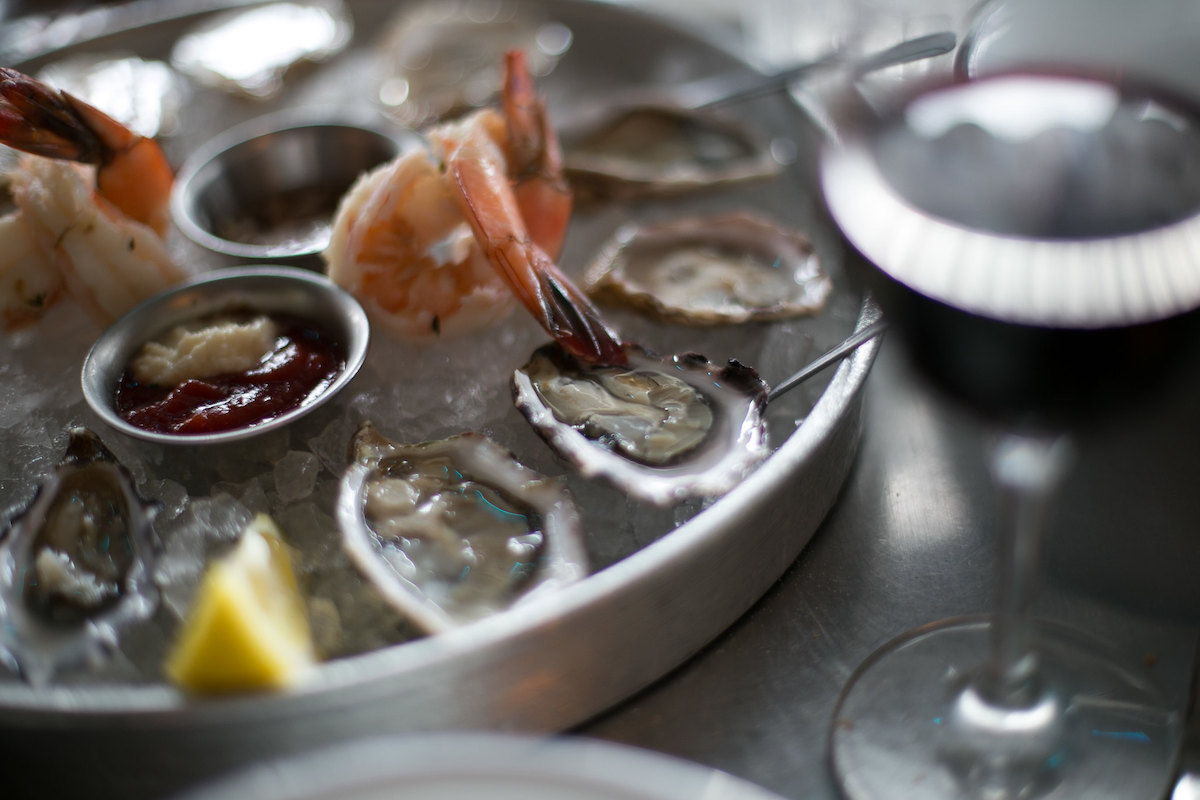 extreme close up of a metal dish with oysters on ice, metal tins of sauce, and cooked shrimp. A glass of red wine is blurred in the foreground.