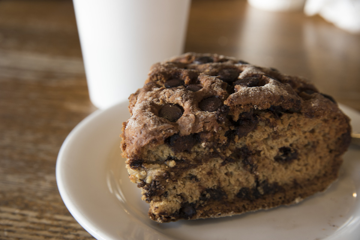 Close up of a chocolate chip scone with a paper cup full of coffee in the background.