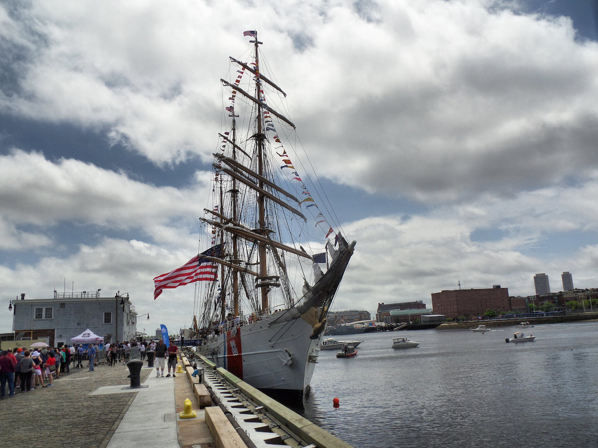 People line up on the pier to board a tall ship flying an American flag