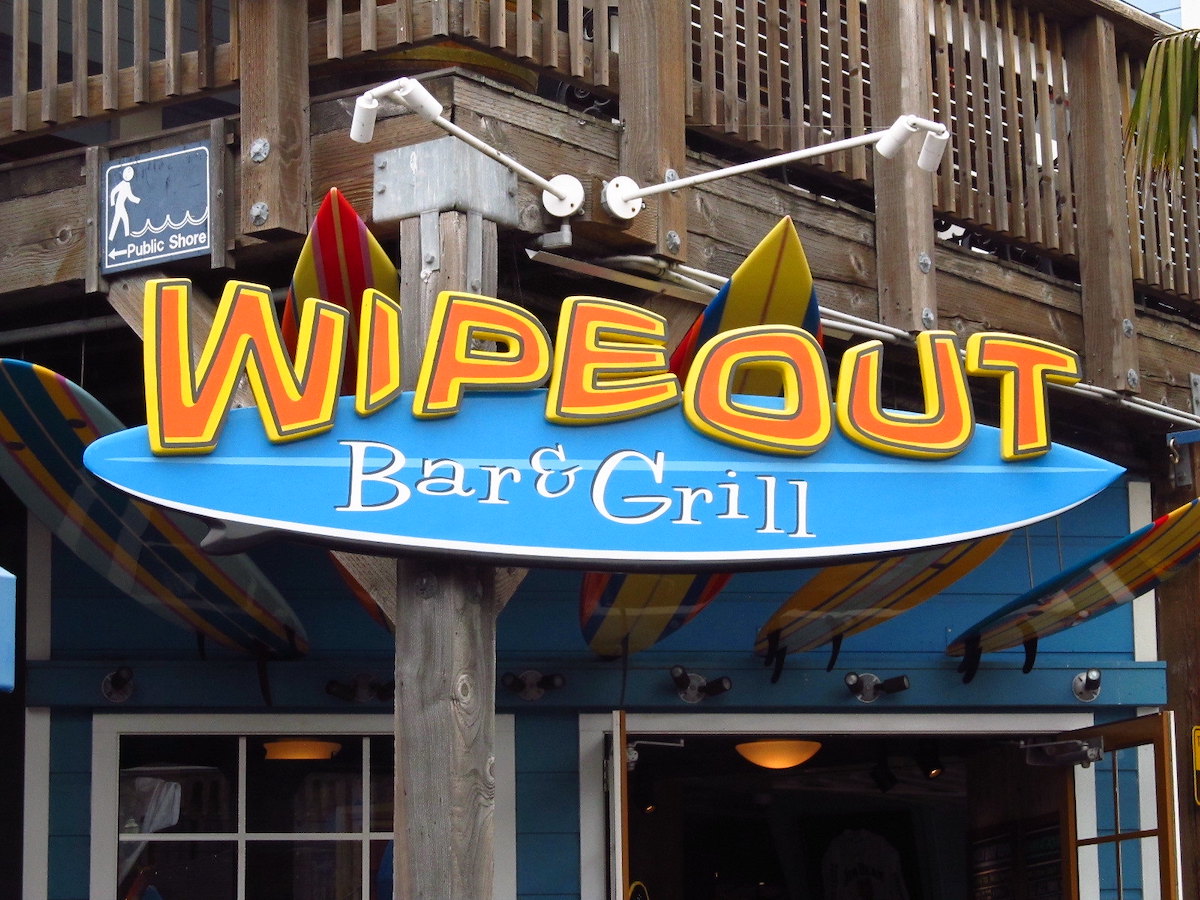 Photo of a restaurant sign with "Wipeout" in big orange and yellow letters atop a bright blue surfboard shape. Wipeout Bar & Grill is a restaurant on San Francisco Pier 39.