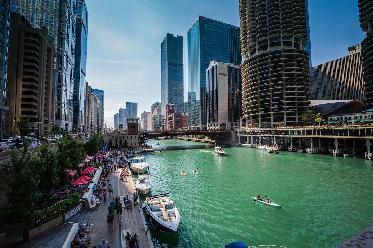 small boats and people in canoes float in the Chicago river while people walk along the riverwalk