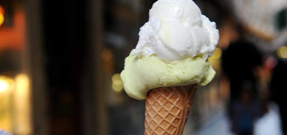 close up of a person's hand holding a small waffle cone with two scoops of gelato, one light green and one white, with a Venice street in the background