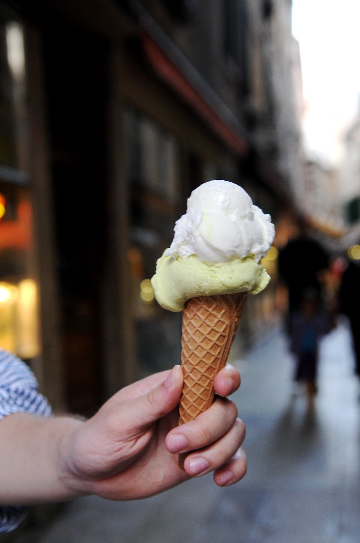 close up of a person's hand holding a small waffle cone with two scoops of gelato, one light green and one white, with a Venice street in the background 