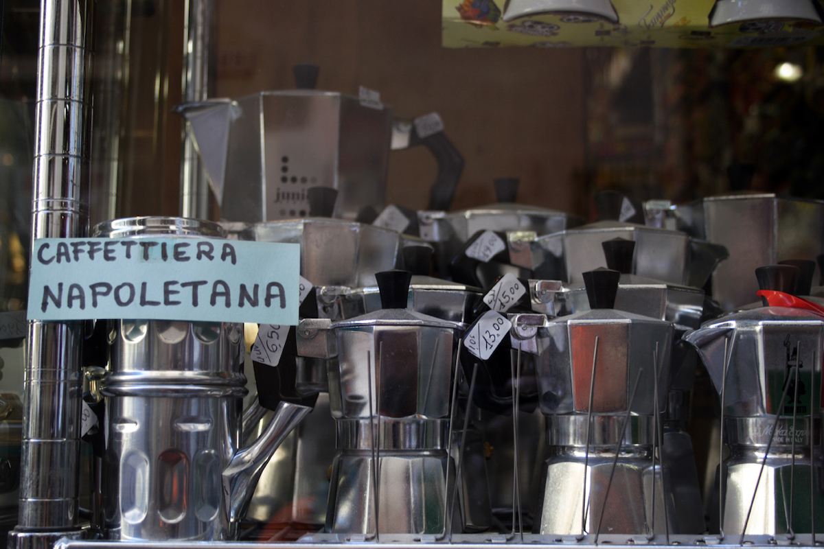 A display of at-home coffee pots in Italy. One Naples style cuccuma pot is on the right with a paper sign that reads "caffeteria napoletana", the rest of the shelf is filled with shiny Moka pots