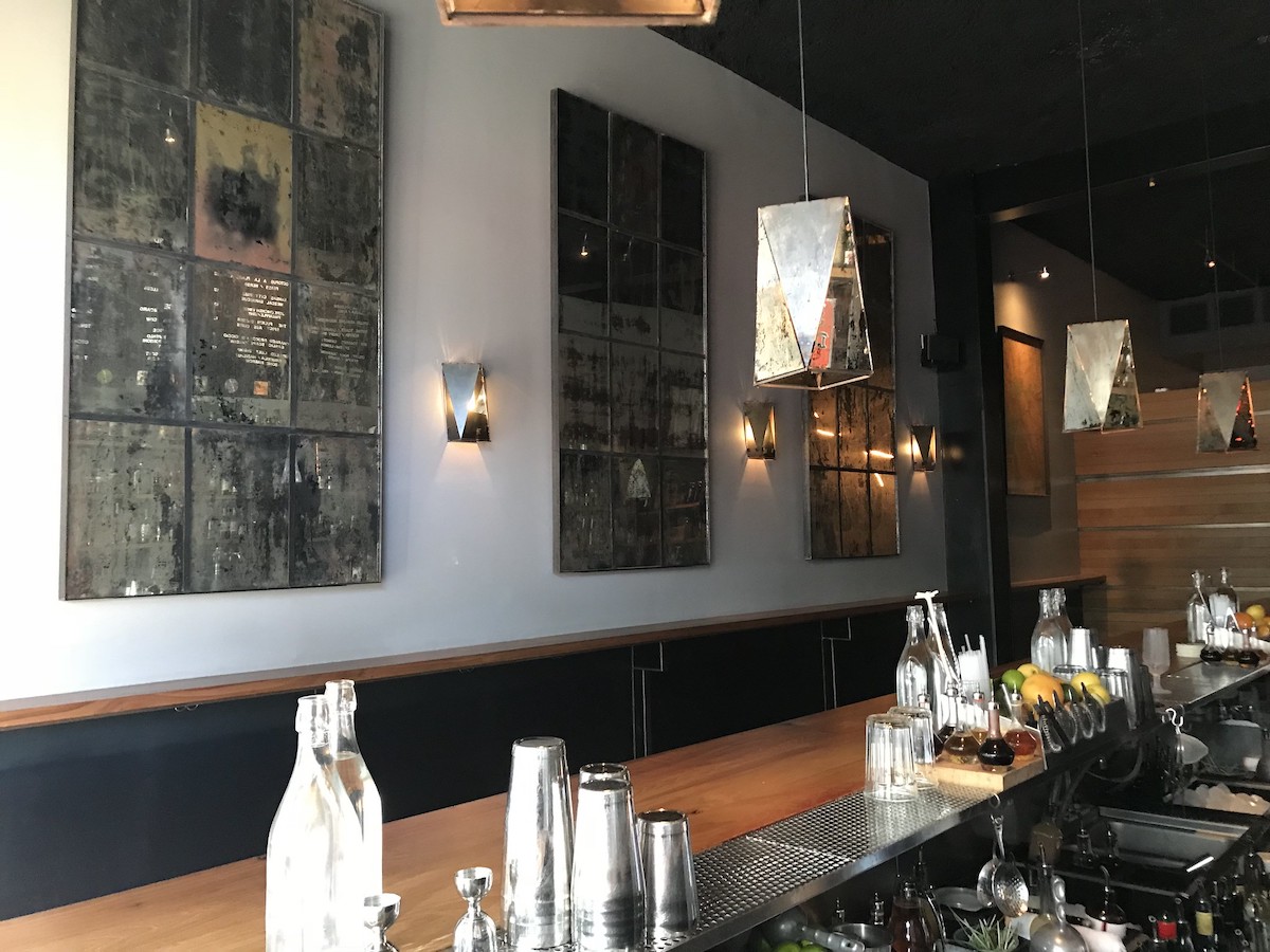 Interior shot from behind the bar of ABV, a cocktail bar in San Francisco. Restored mirror panels decorate the walls, reflecting the shelves of the bar and the menu on the wall
