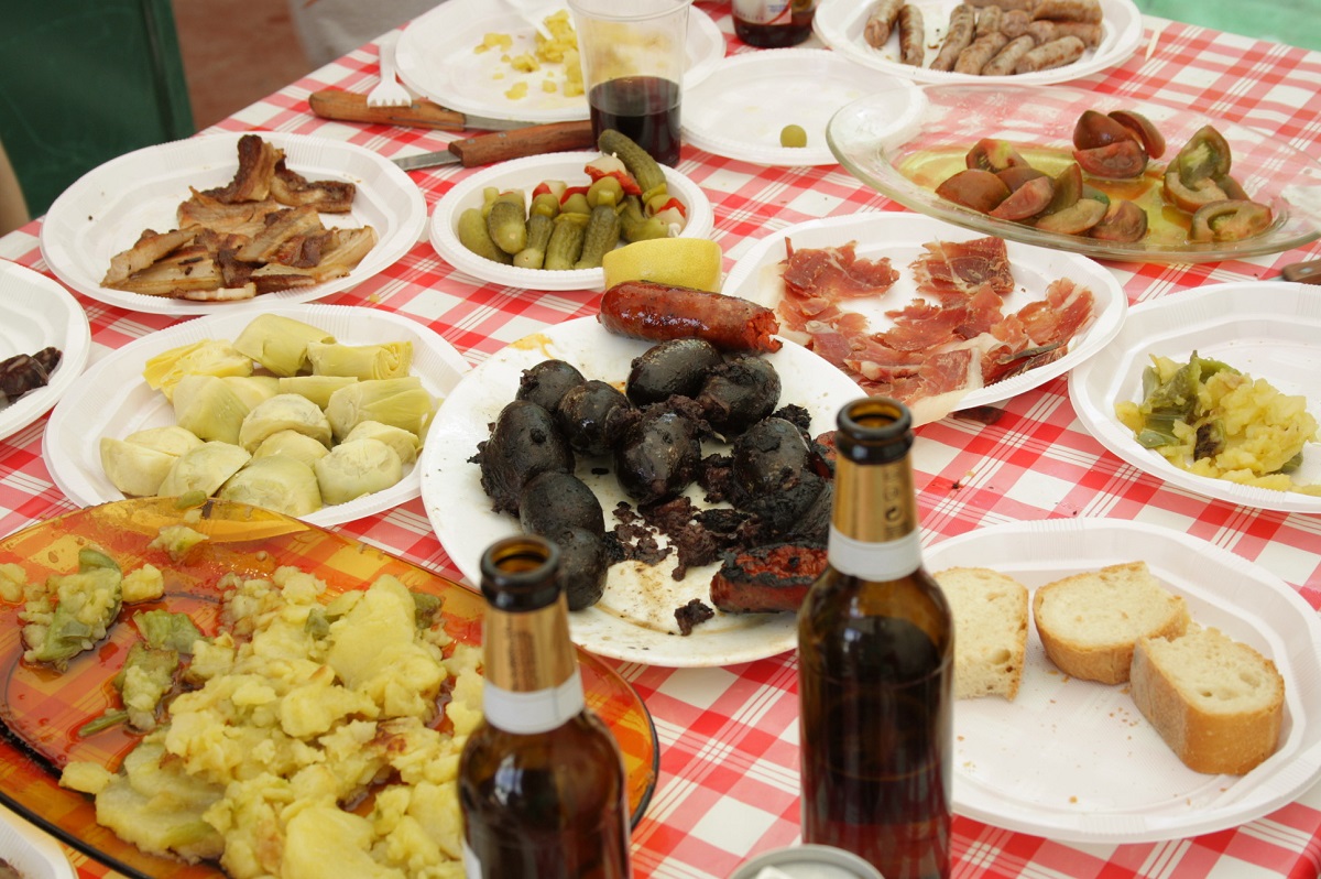 Picnic table spread with lots of small Spanish finger foods such as morcilla (blood sausage), ham, cheese, olives, pickled artichokes, and bread