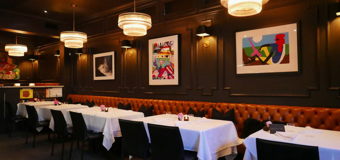Interior shot of the dining room of Californios, one of the best restaurants in San Francisco. Tables with white tablecloths line up along a long brown leather booth. The walls are painted black and colorful abstract artwork hangs on the walls.