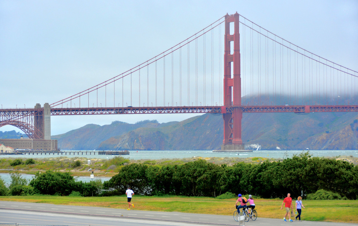 People jog, walk, and ride bikes along a path at Crissy Field in San Francisco with the Golden Gate Bridge towering in the background.