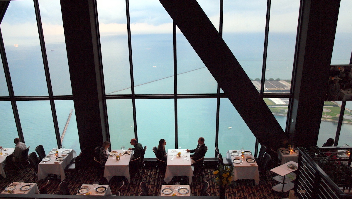 Shot from the interior of the Signature Room in Chicago, with floor to ceiling windows looking out on the water. People sit at tables with white tablecloths and enjoy their food.
