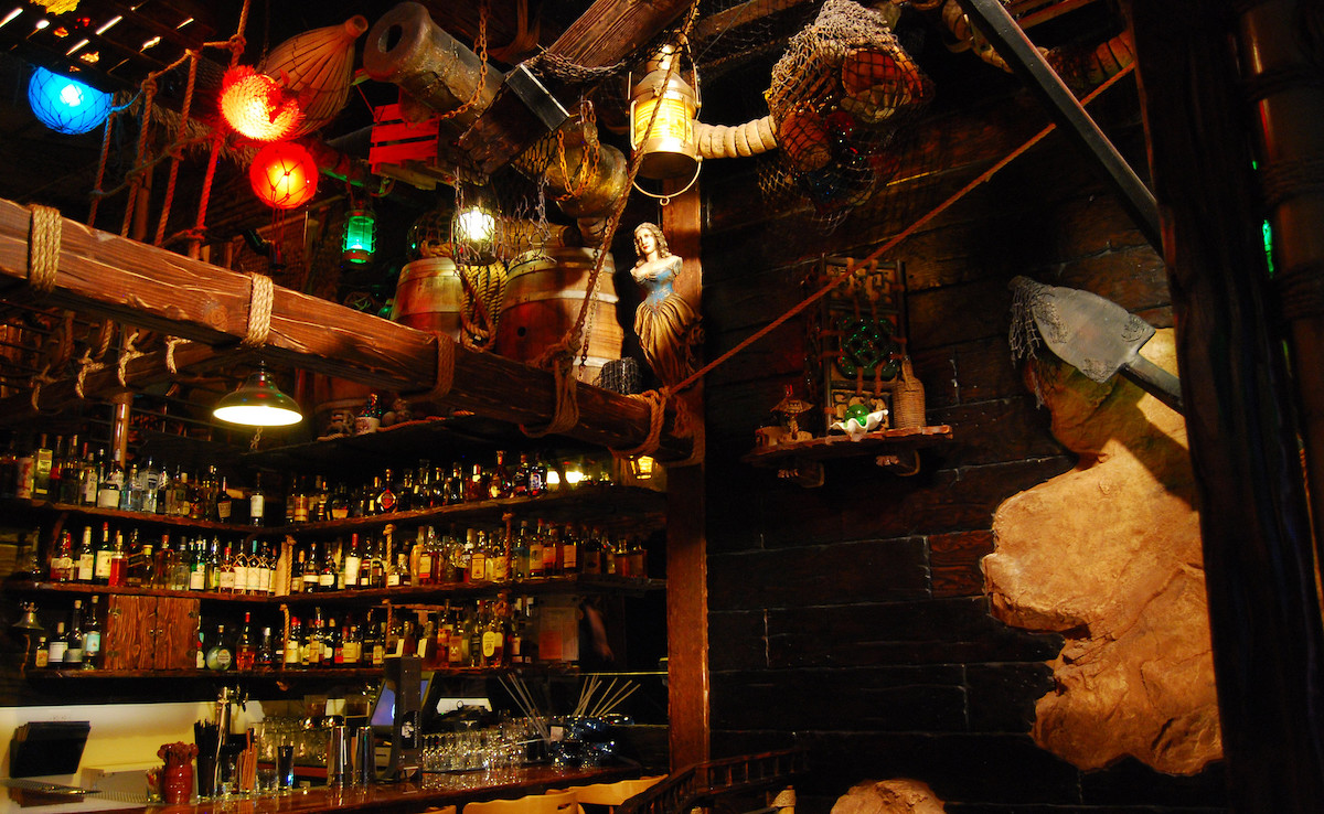 Interior of Smuggler's Cove tiki cocktail bar in San Francisco. Dark wood paneling surrounds the space with shelves full of liquor bottles behind the bar.