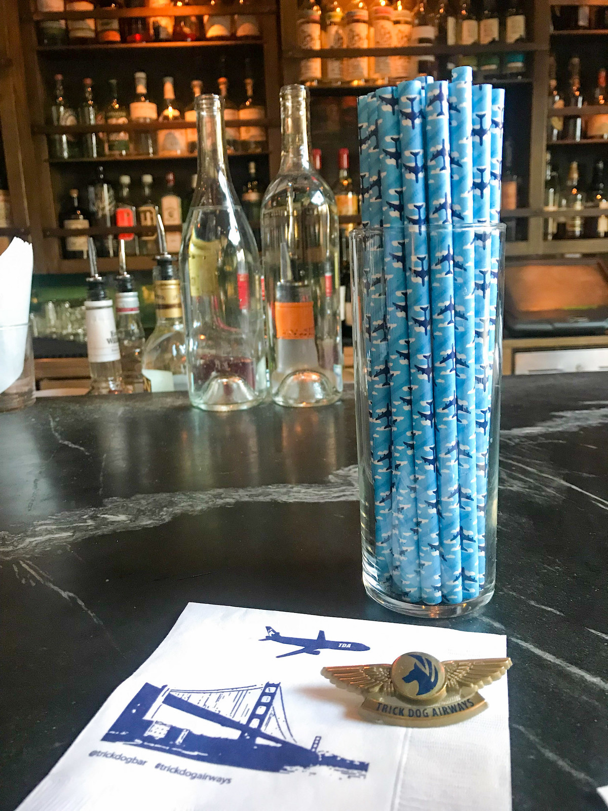 Picture of the marble bar at Trick Dog Bar in San Francisco, with a glass full of blue straws decorated with airplanes and shelves of liquor bottles behind the bar
