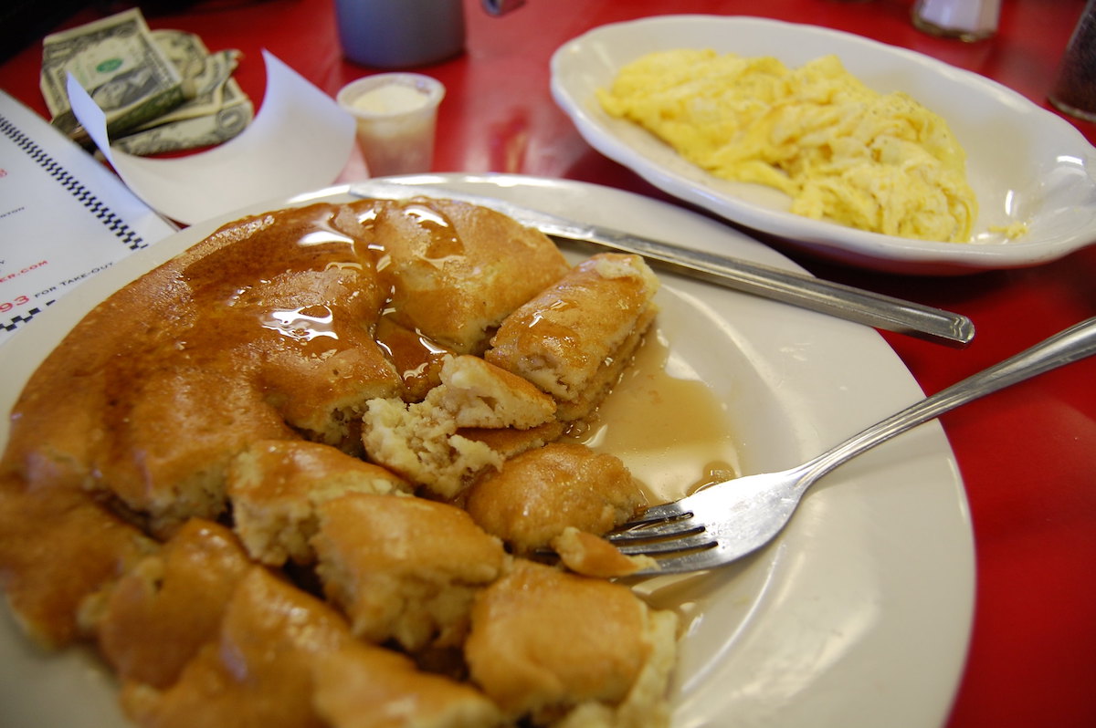close up of a red table with two white plates, one with pancakes that have been cut drizzled with syrup, the other with yellow scrambled eggs