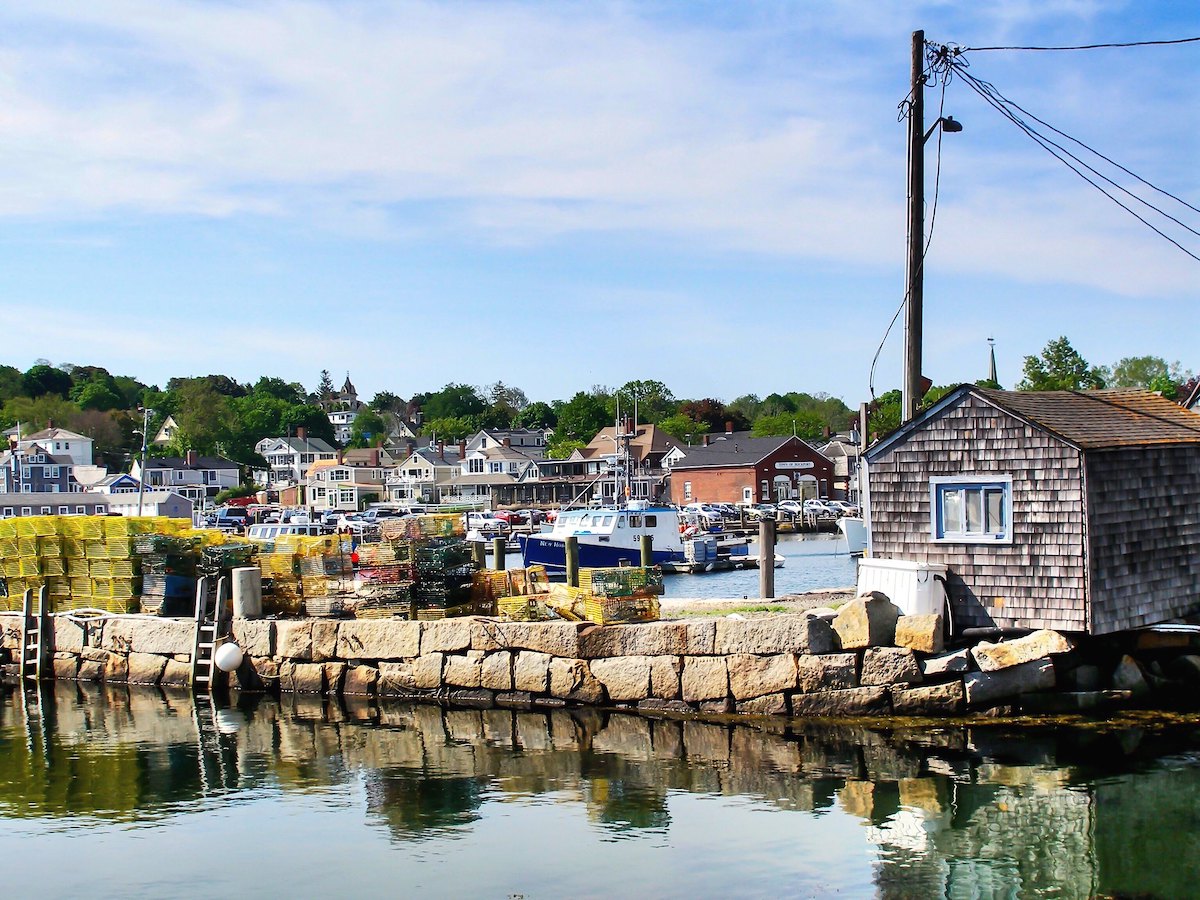 Crates are piled up on a dock near the water of a bay in Rockport, Massachusetts. Boats float in the background