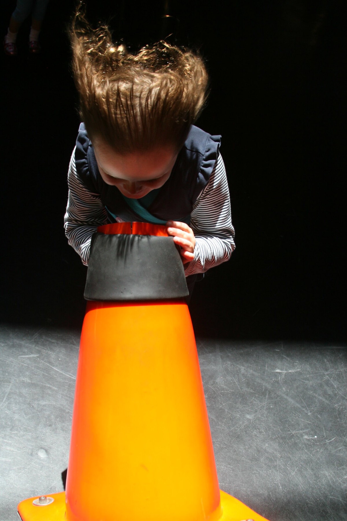 A child stands with their face above an orange cone, with a wind blowing their hair back.