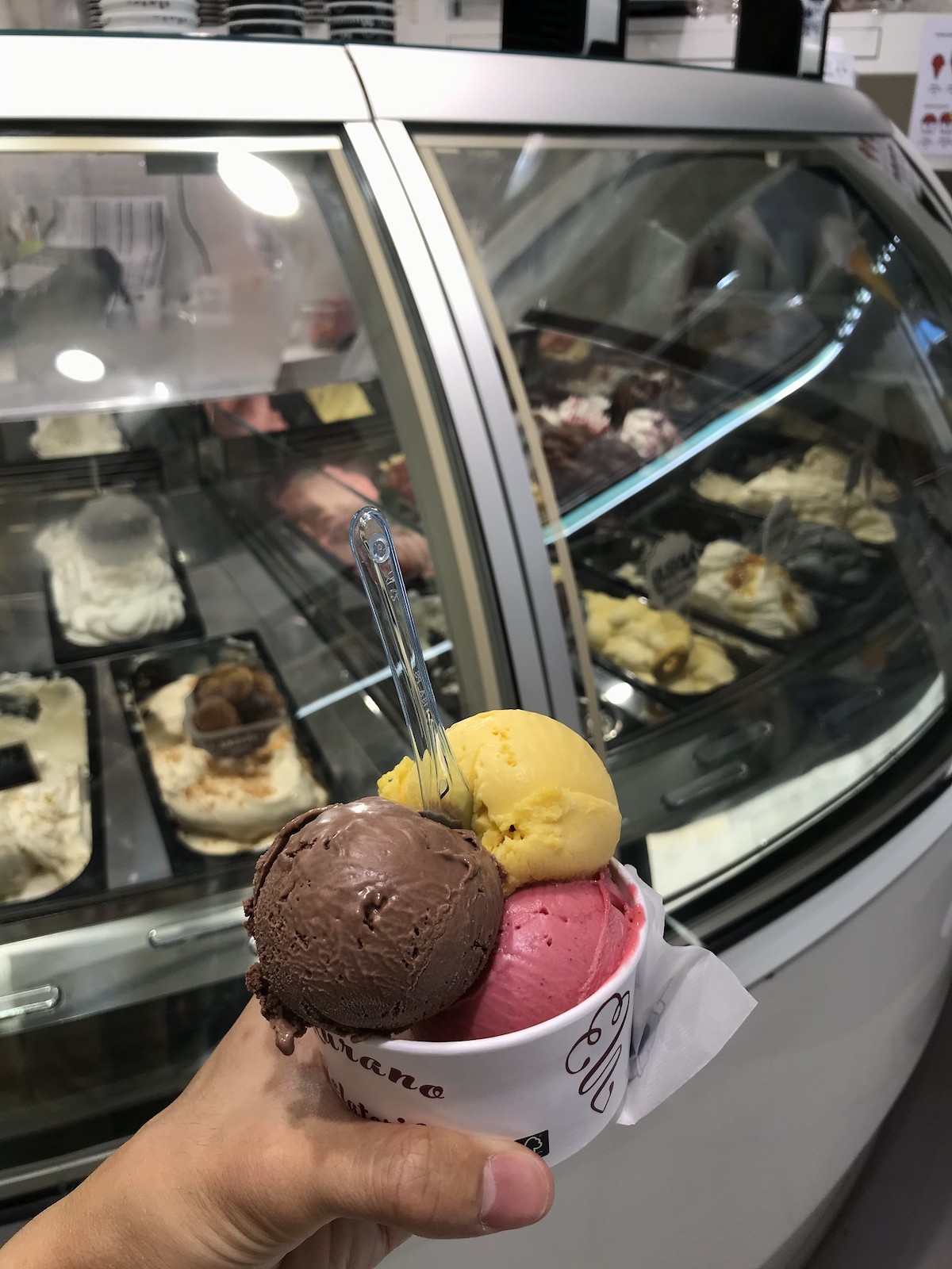 A person holds a white cup with three scoops of gelato, one brown, one yellow, one pink, at a gelato shop in Venice