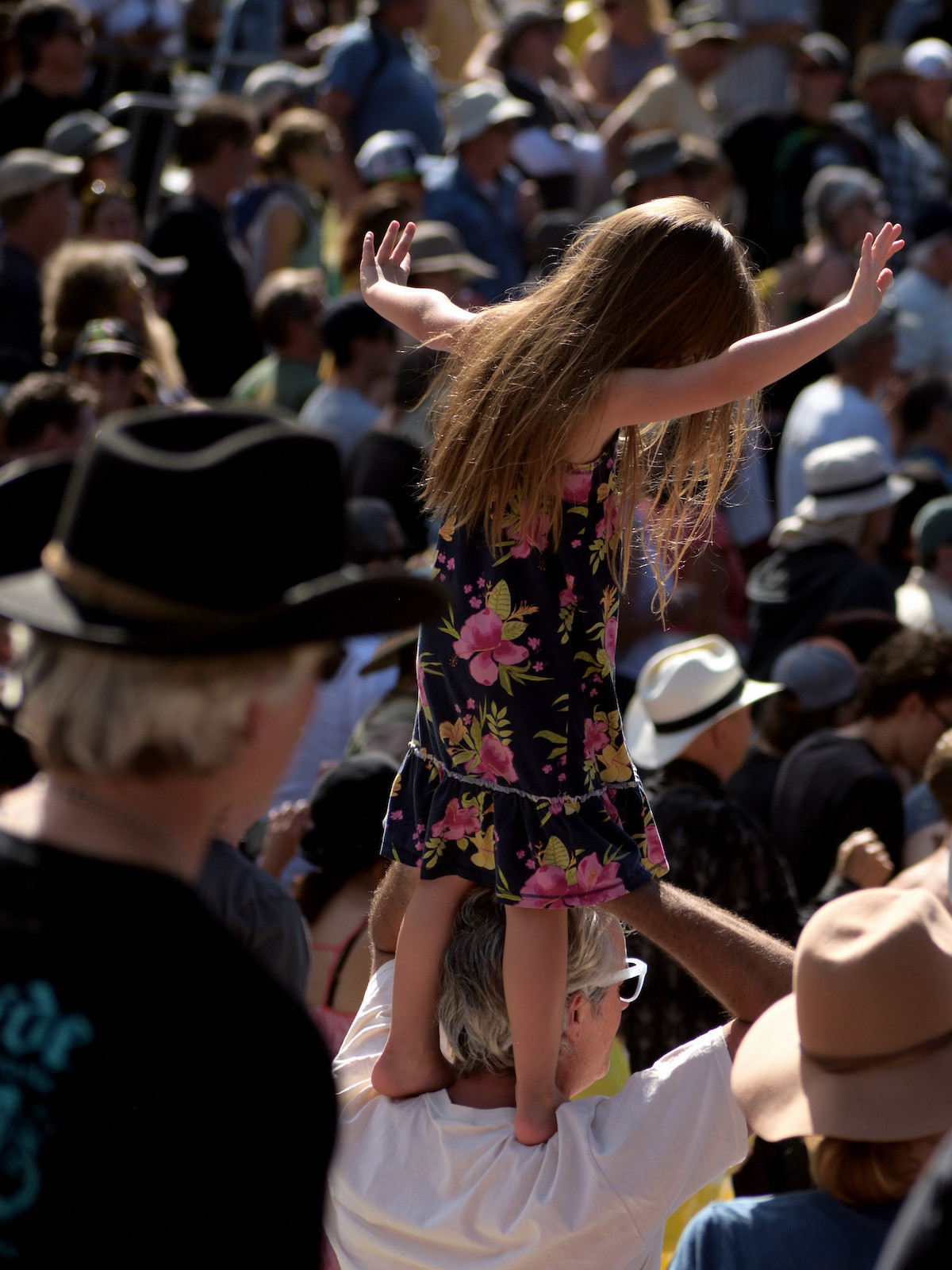 A girl wearing a black dress with pink flowers stands on a man's shoulders in a large crowd at a music festival in summer in San Francisco