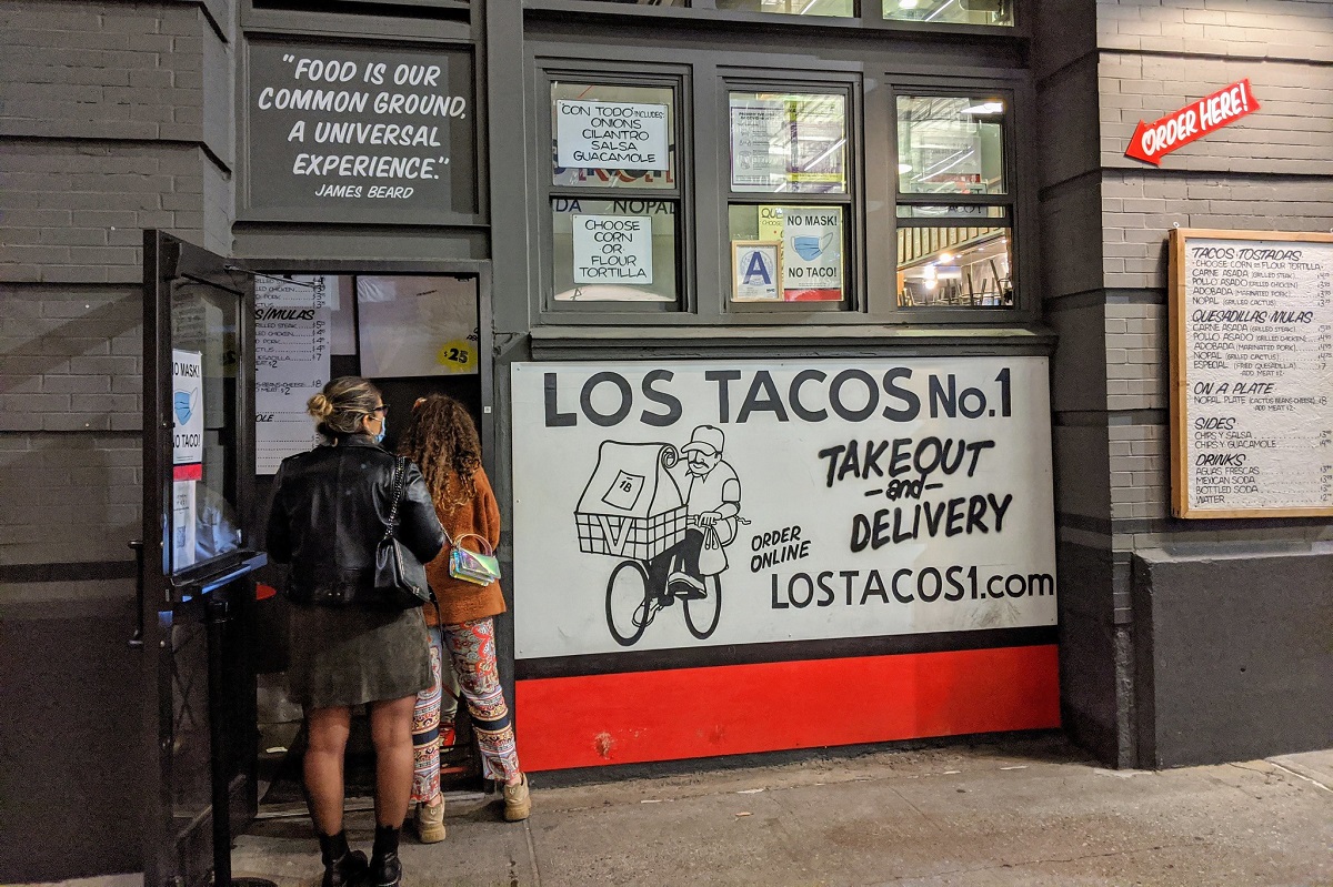 People line up for food outside of a grey colored taco stand