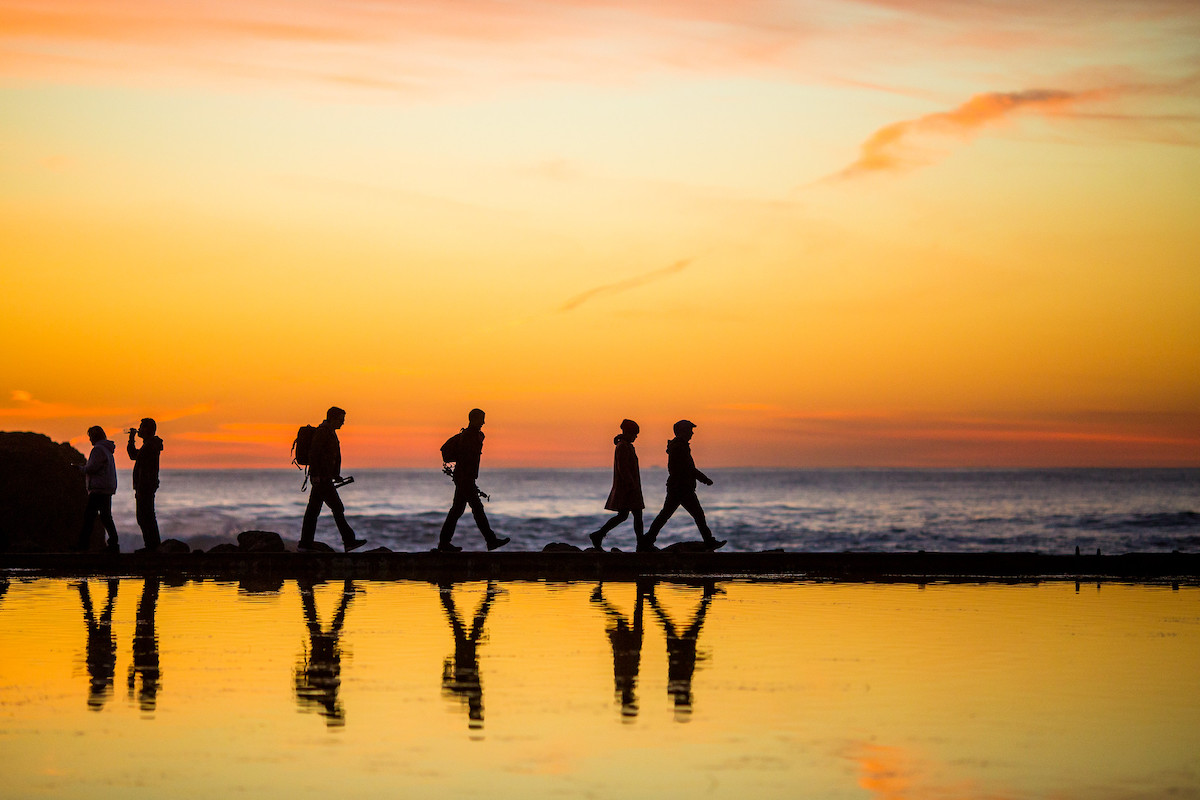 The silouhettes of four people walking is reflected in the water of the Sutro baths, the sunset sky is golden orange