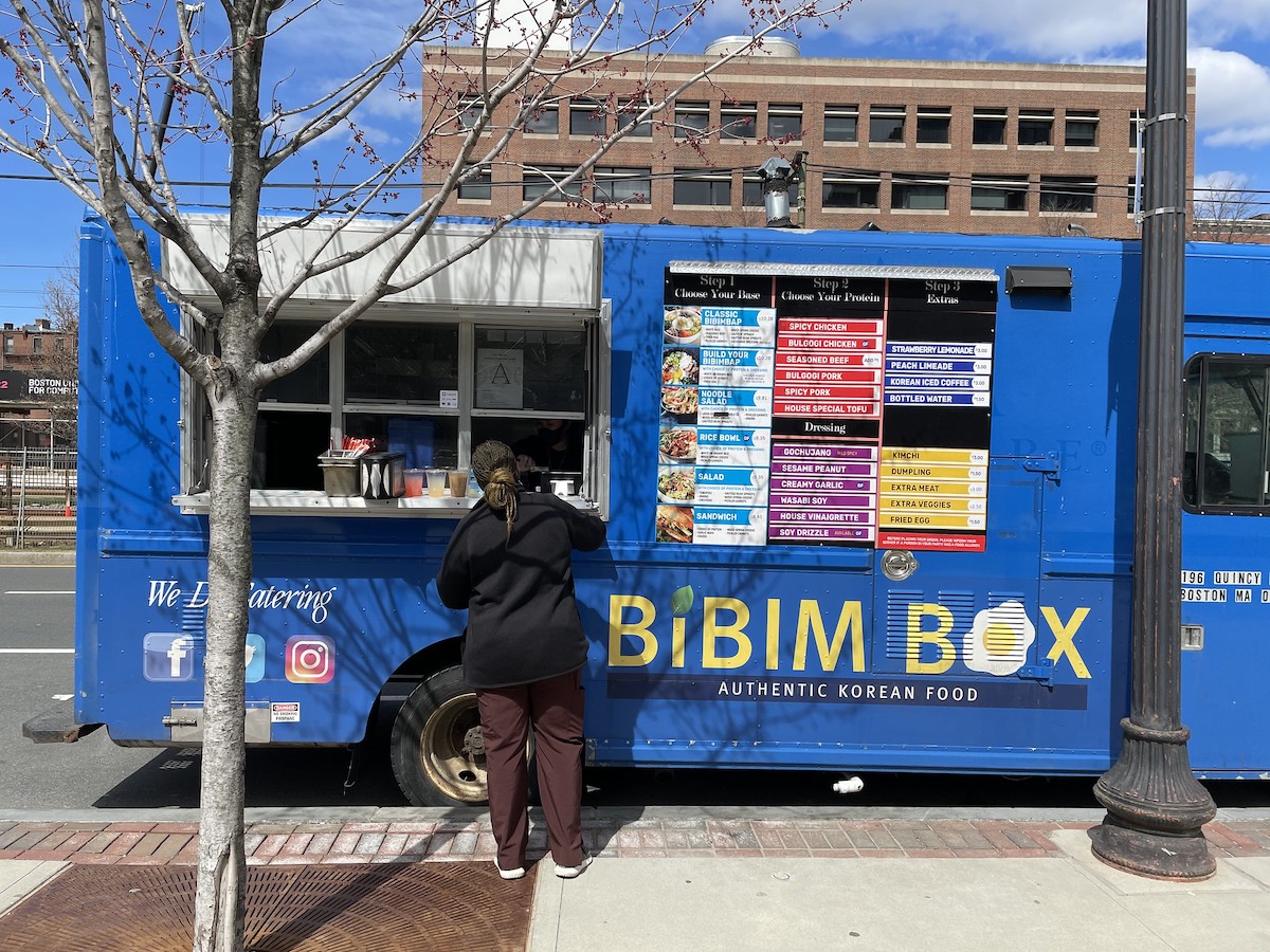 a person stands at the order window of a blue food truck in Boston. Bright yellow letters read "Bibim Box" on the side of the truck.