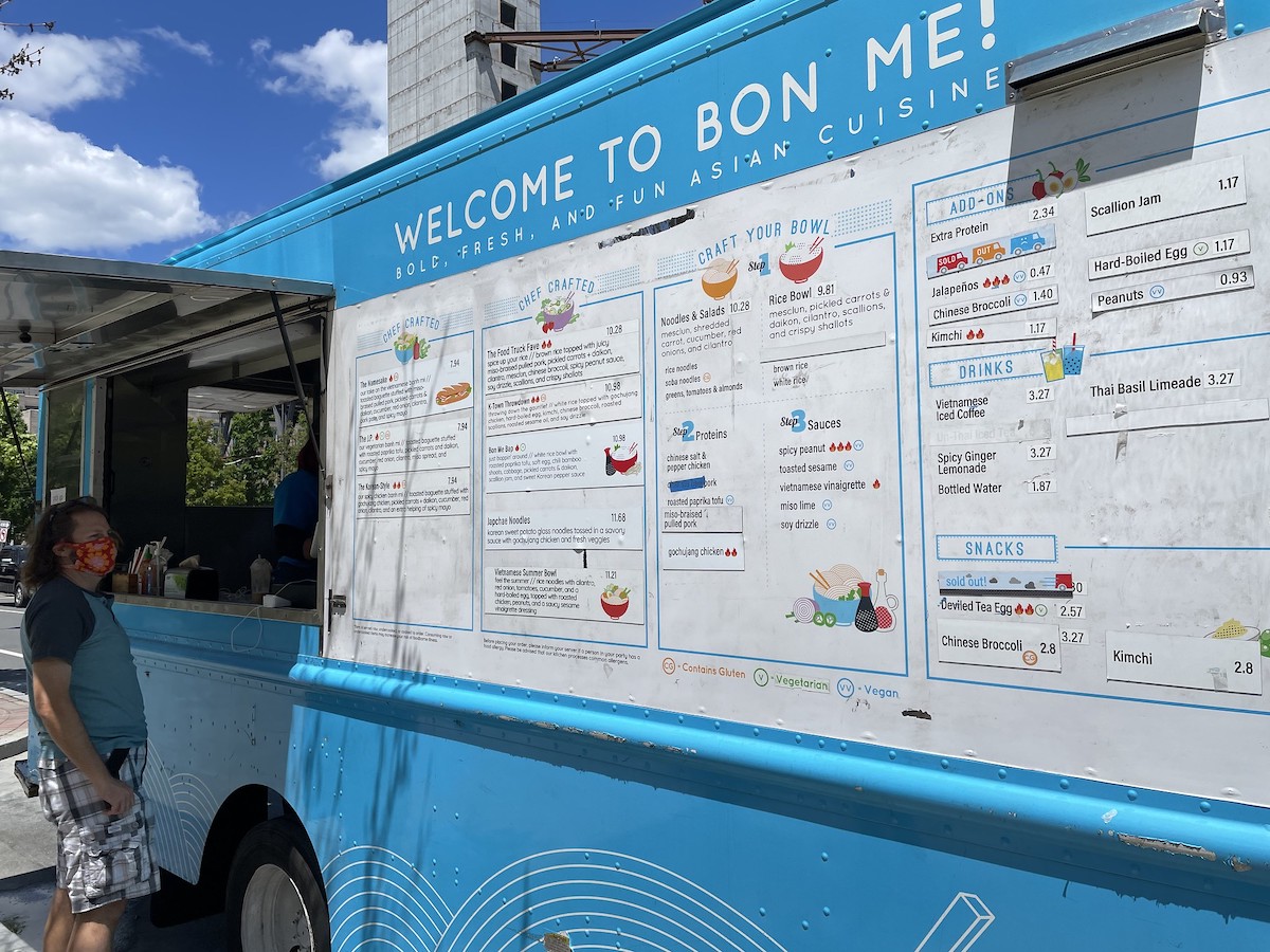 A person stands at the order window of a blue food truck in Boston. A large, white menu with colorful images stretches across the side of the truck. 