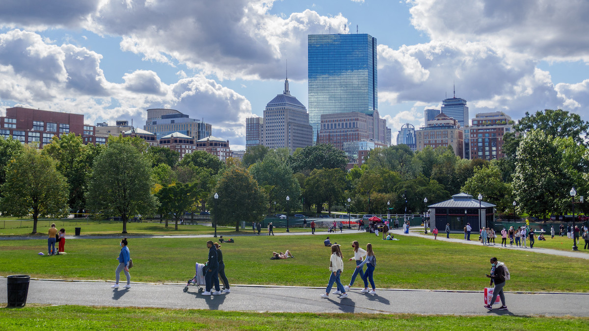 Wide shot of Boston Common, people walk on the side walk, some people are laying in the grass, skyscrapers in the background