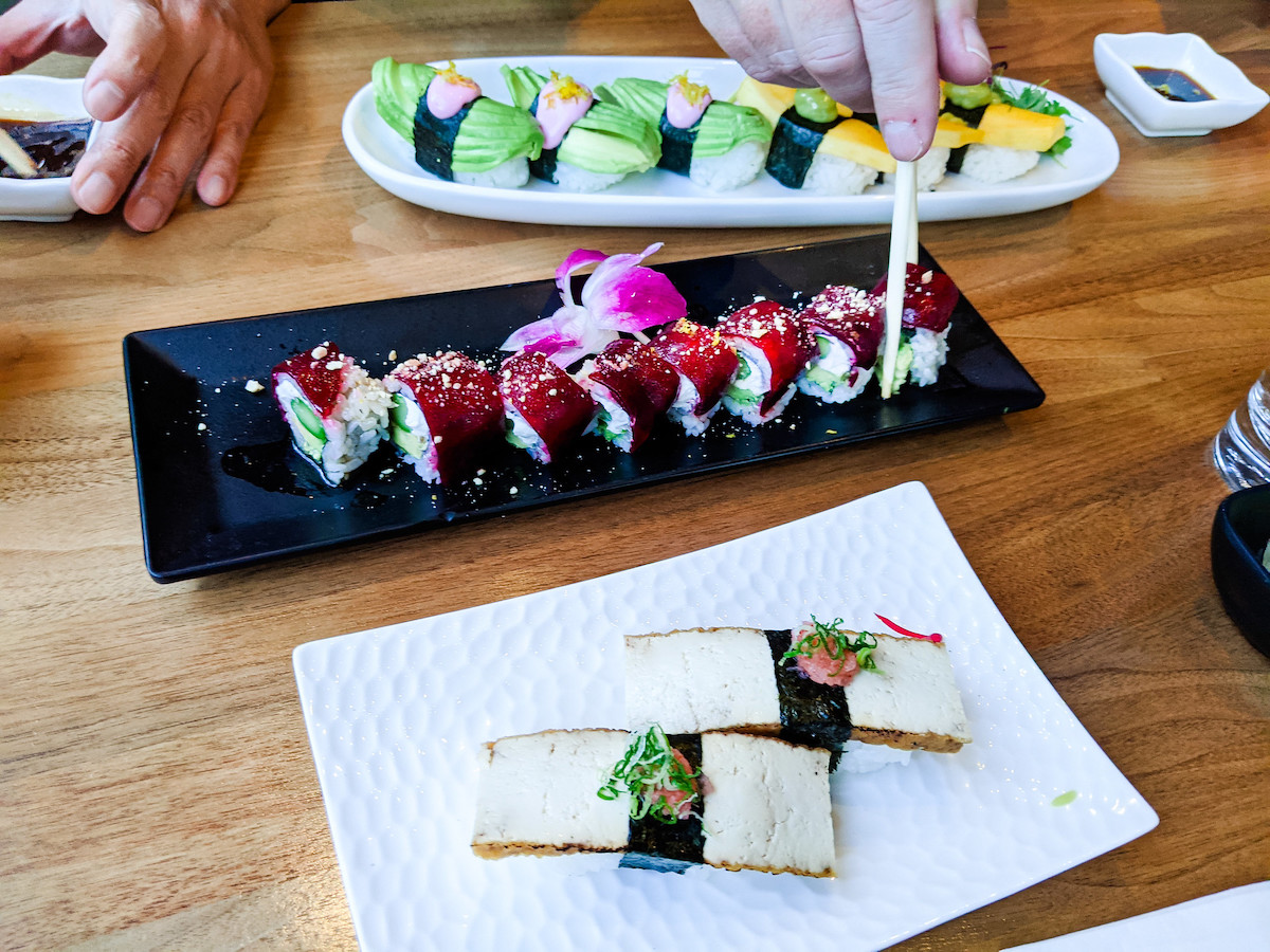 Three plates of sushi sit atop a wooden table, a person uses chosticks to pick up a piece of sushi